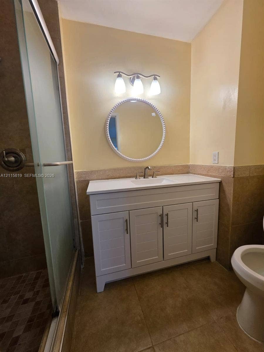831 NE 207th Lane, #2-203, Miami, Miami, FL 33179 White vanity with rectangular undermount sink, circular framed mirror, three-light wall fixture, and glass-enclosed shower with stone tile flooring