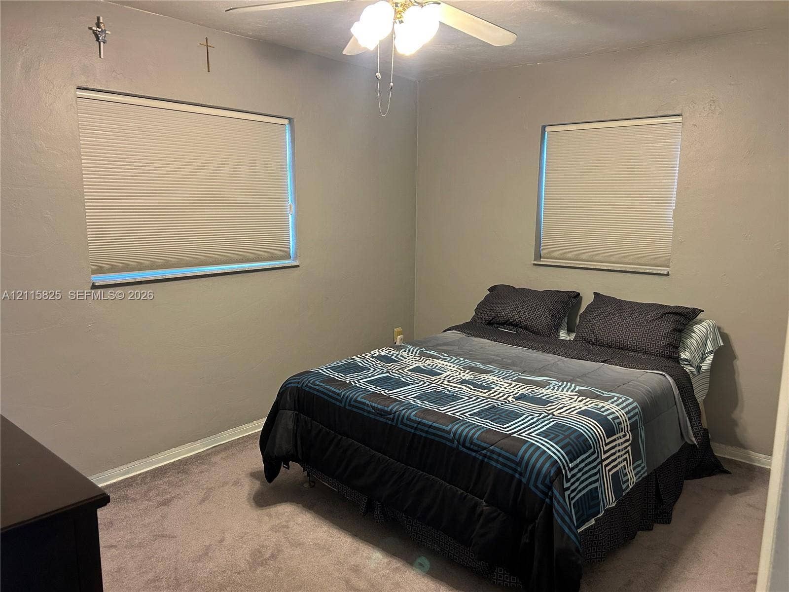 7130 W 12th Court, Hialeah, FL 33014 Neutral-toned interior room featuring wall-to-wall carpeting, dual windows with pleated shades, and a central ceiling fan with integrated lighting