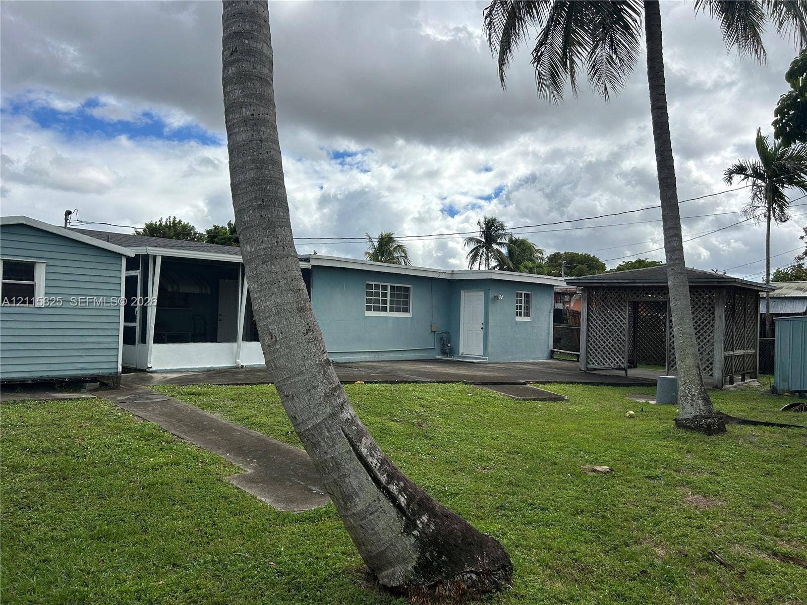 7130 W 12th Court, Hialeah, FL 33014 Exterior featuring light blue lap siding, a screened-in porch, a detached wood-lattice gazebo, and expansive lawn area with mature palm trees