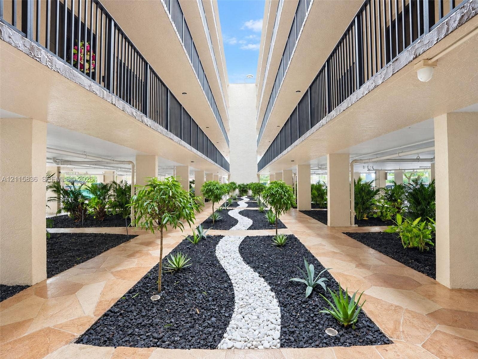 8000 Hampton Boulevard, #311, North Lauderdale, North Lauderdale, FL 33068 Multi-story building featuring structural columns, decorative black metal railings, stone-paver ground surfaces, and manicured landscaping with organic rock borders