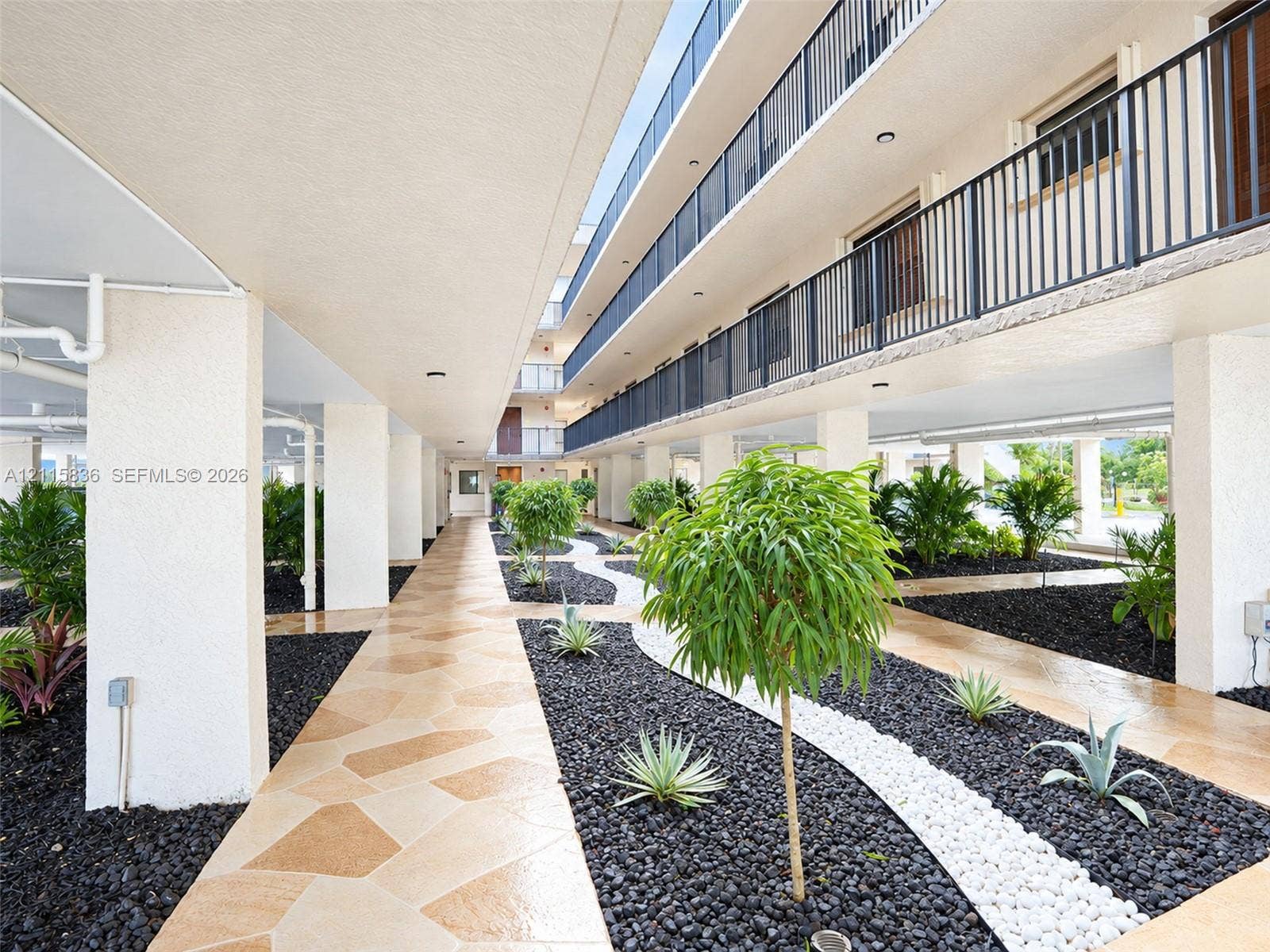 8000 Hampton Boulevard, #311, North Lauderdale, North Lauderdale, FL 33068 Covered ground-level walkway featuring paver stone pathways, dark volcanic rock landscaping, white decorative stone borders, integrated ornamental shrubbery, and structural concrete pillars