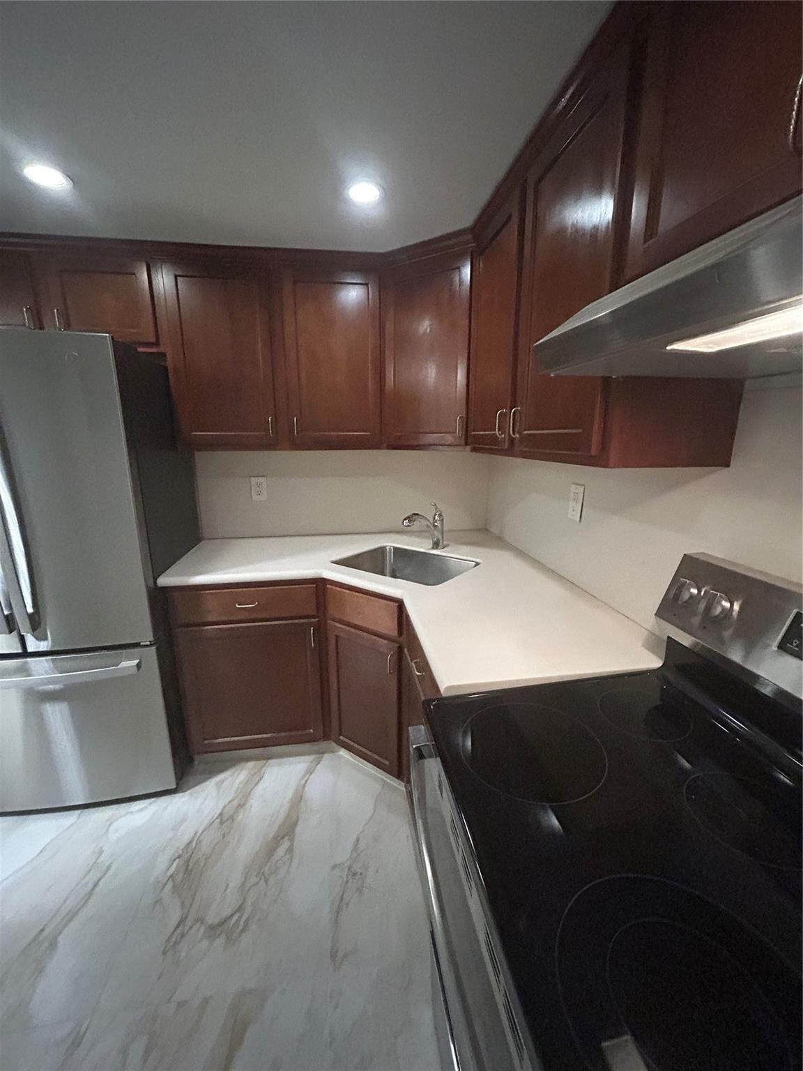 550 Bayshore Drive, #102, Fort Lauderdale, FL 33304 recessed lighting from a dimmer switch shine on wood cabinets above and below. New appliances include refrigerator, stove and hood. White marble counter tops with steel sink and hosed faucet have lights under cabinets. Steel appliances on ceramic tile with similar vein colored flooring.
