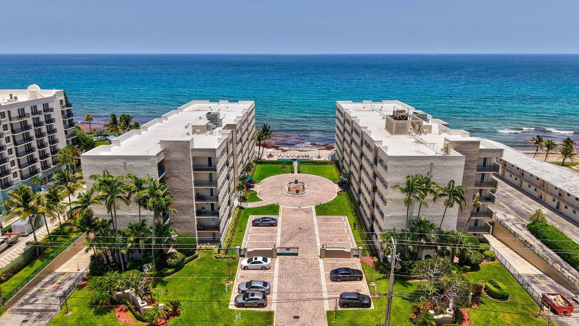 1239 Hillsboro Mile, #302, Hillsboro Beach, FL 33062 Oceanfront apartment buildings featuring private balconies, mature palm trees, manicured lawns, and a paver drive with central fountain