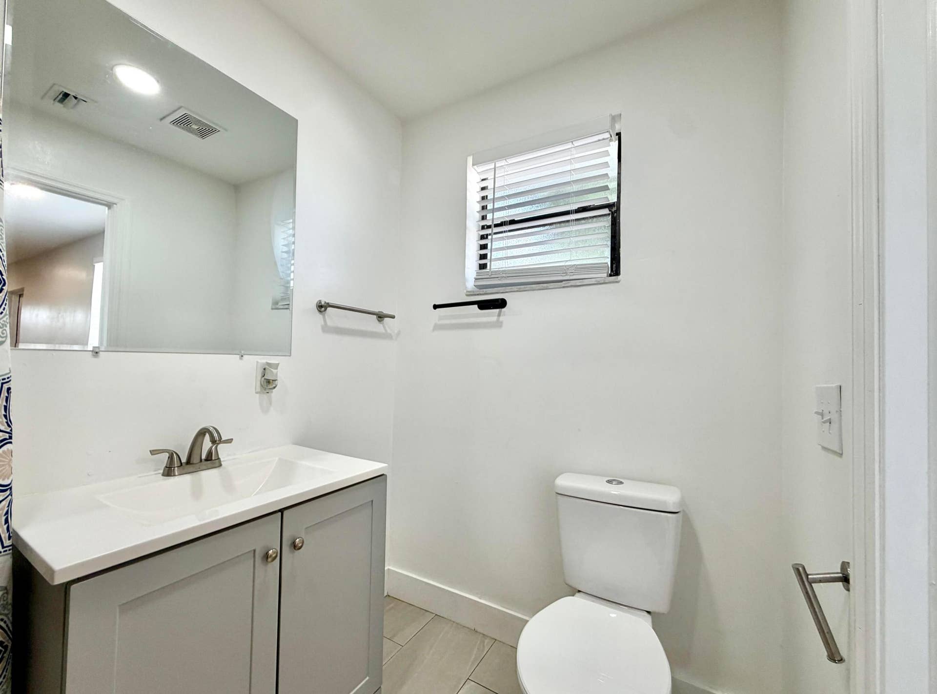 Bathroom featuring a light gray vanity with a white countertop and integrated sink, brushed nickel faucet, large wall-mounted mirror, and horizontal window blinds