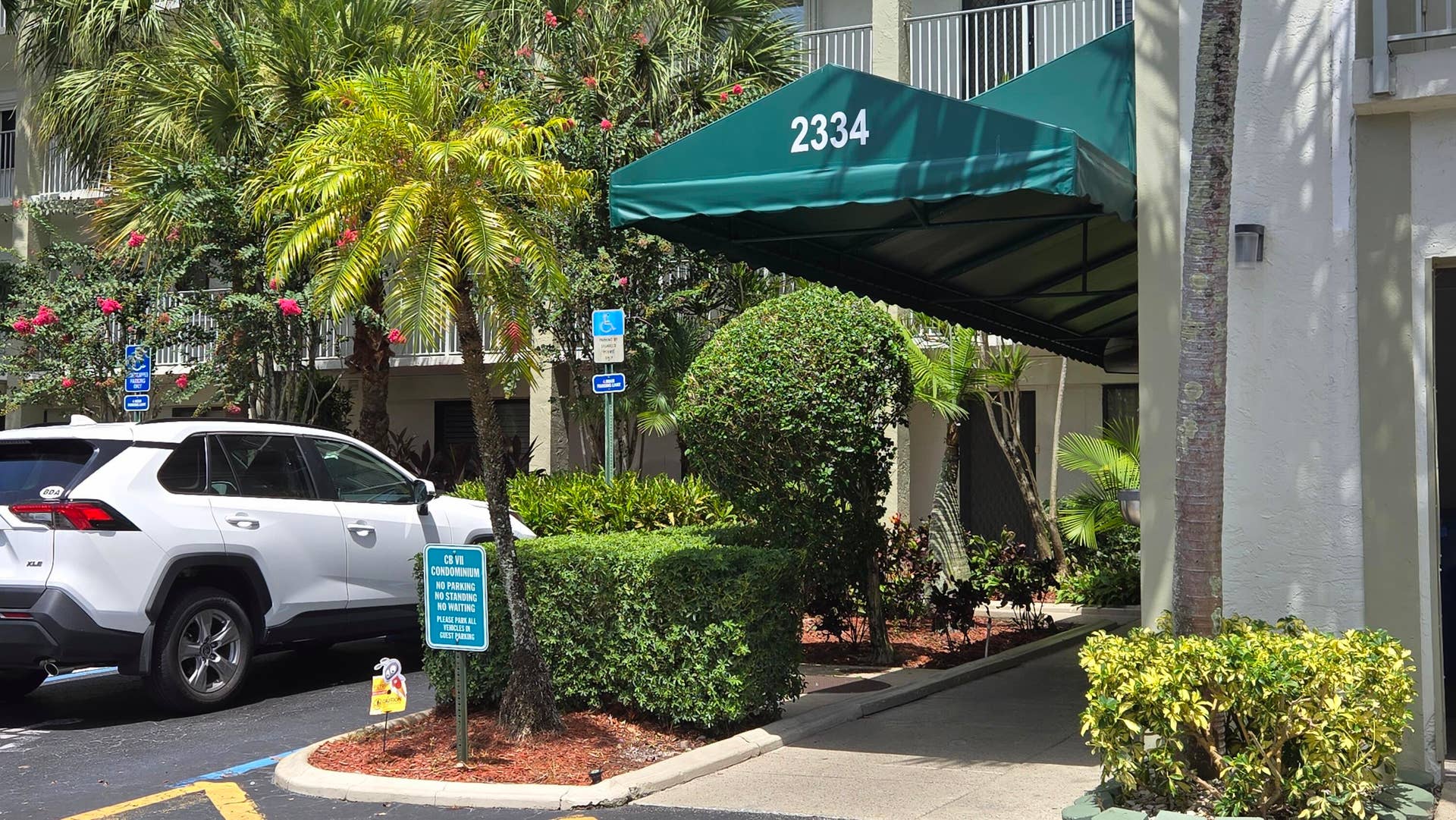 2334 S Cypress Bend Drive, #602, Pompano Beach, FL 33069 Building entrance featuring a green awning, surrounding lush tropical landscaping with palm trees and manicured hedges, and a dedicated drop-off area