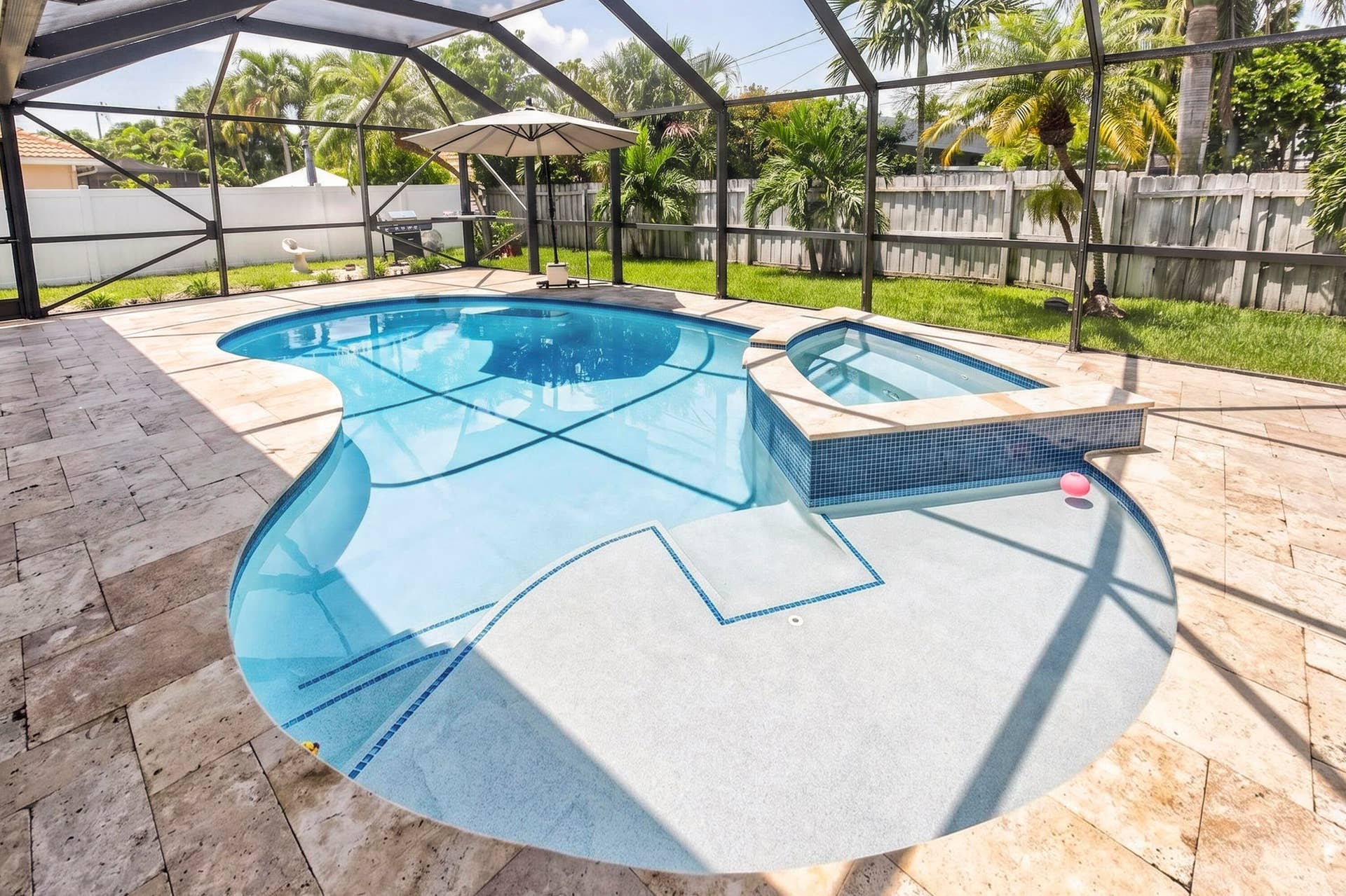 Screened outdoor area featuring a freeform swimming pool with integrated spa, light-colored travertine tile decking, and a fenced perimeter