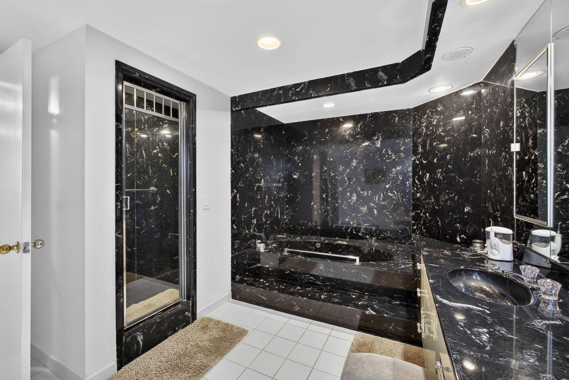3140 S Ocean Boulevard, #101 S, Palm Beach, FL 33480 Bathroom featuring black patterned wall panels, a built-in tub with matching surround, a glass-enclosed shower, a vanity with a black integrated sink, and white floor tiles