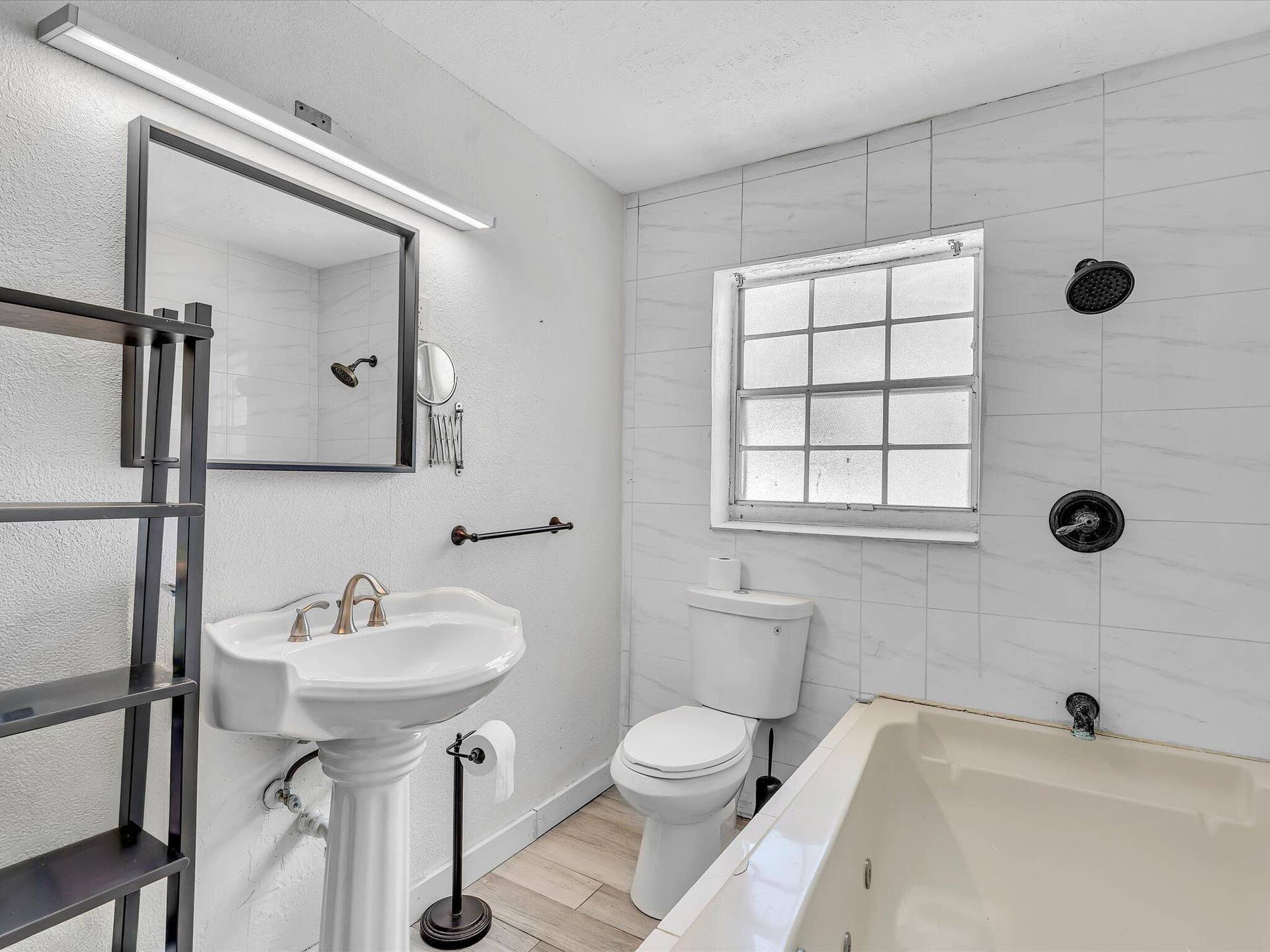 Bathroom featuring a pedestal sink, framed mirror with integrated lighting, tiled walls, a window with grid panes, and a built-in tub with black fixtures