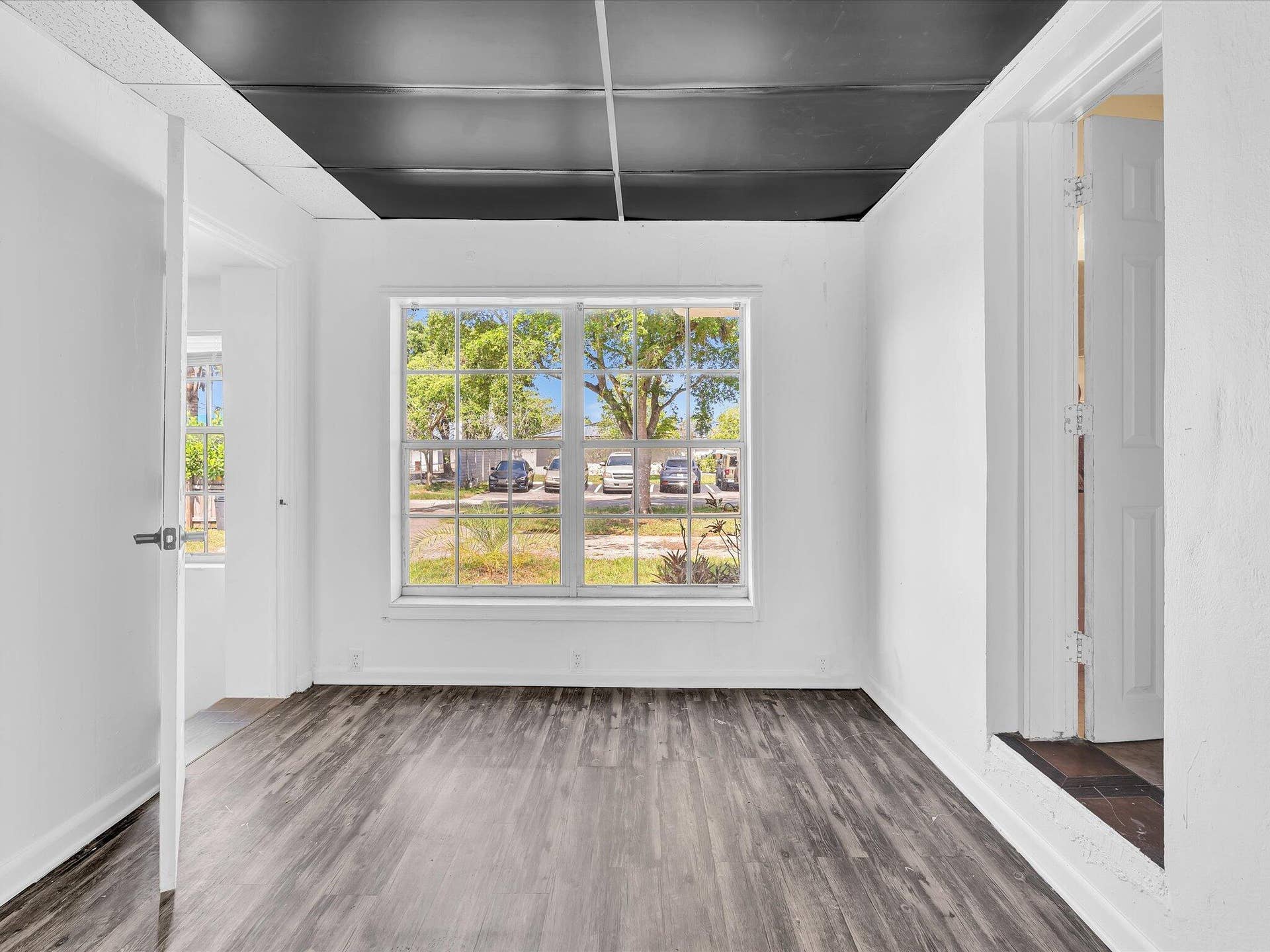 Wood-finish flooring throughout, featuring a large multi-pane window, white painted walls, and a dark ceiling