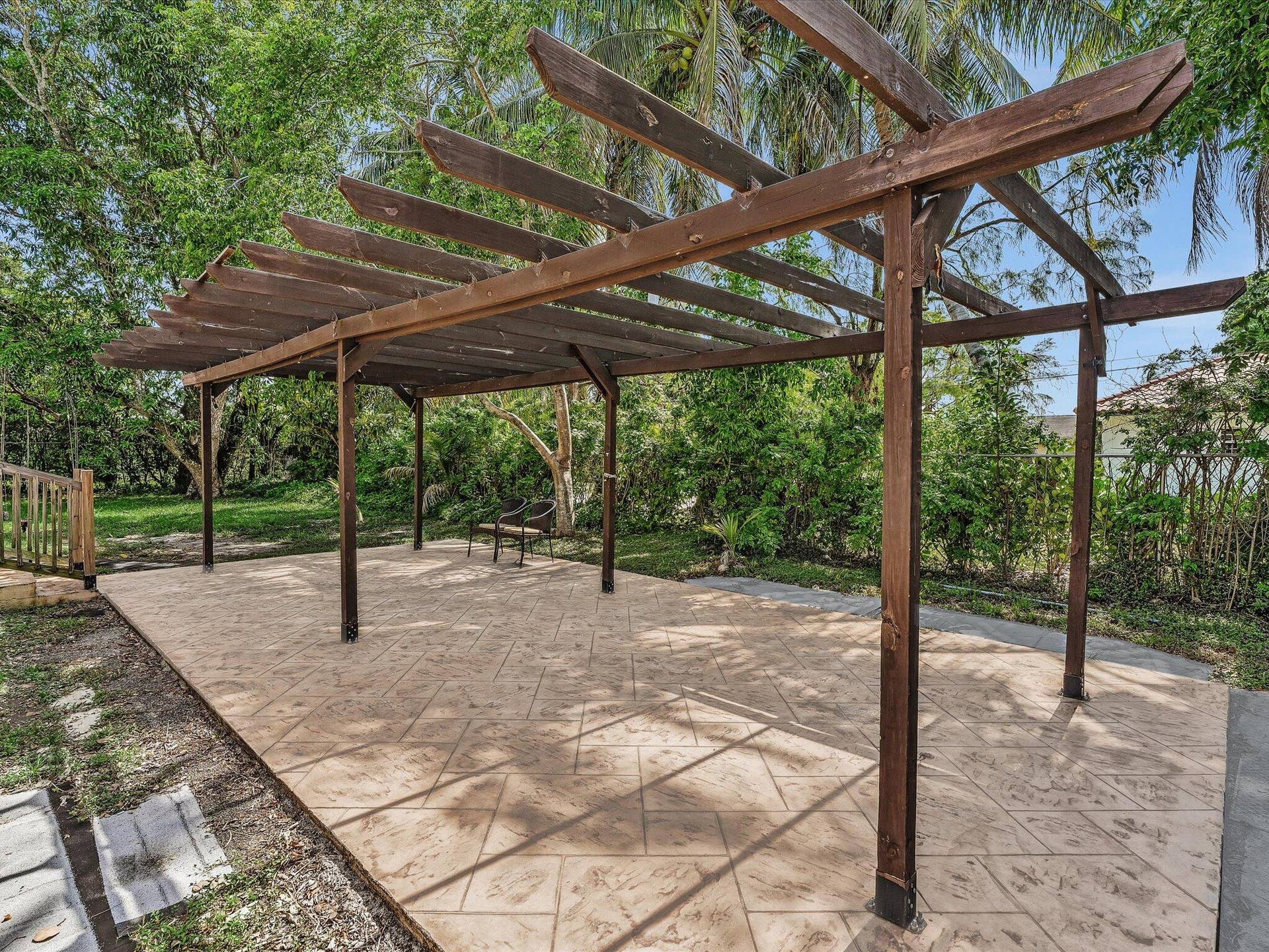 Expansive outdoor patio featuring a large wooden pergola, stamped concrete flooring, lush green landscaping, and mature trees