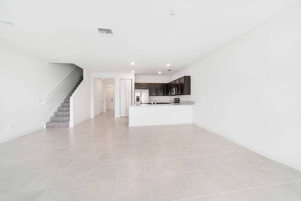 Open-concept living area featuring light-toned tile flooring, a staircase with carpeted treads, and a kitchen with dark cabinetry, light countertops, and stainless steel appliances