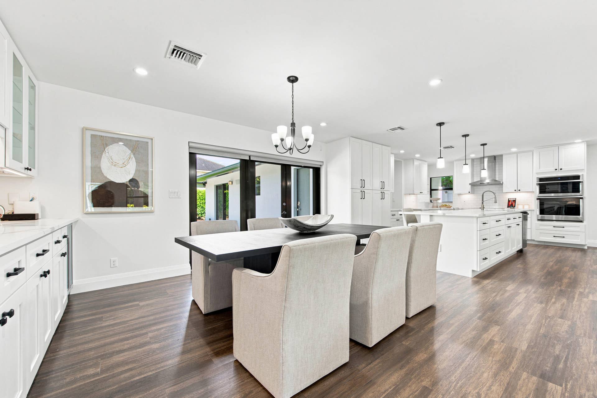 Bright, open-concept kitchen and dining area featuring wood-finish flooring, white cabinetry with dark hardware, a large kitchen island with pendant lighting, stainless steel double wall ovens, and sliding glass doors