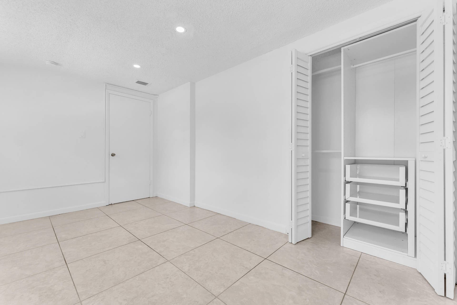 Bright room featuring neutral tile flooring, white walls, recessed lighting, and a closet with louvered bifold doors and built-in shelving