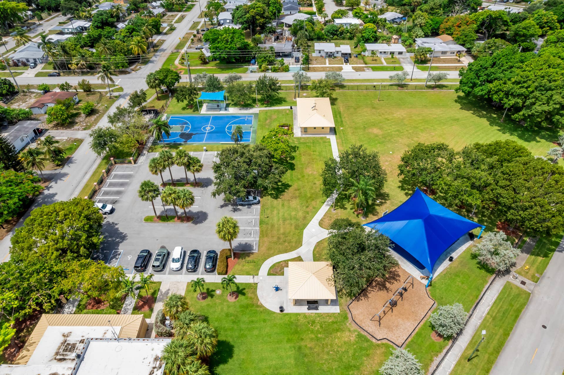 Expansive community park featuring a vibrant blue basketball court, shaded playground area, multiple paved walking paths, open green spaces, and ample parking