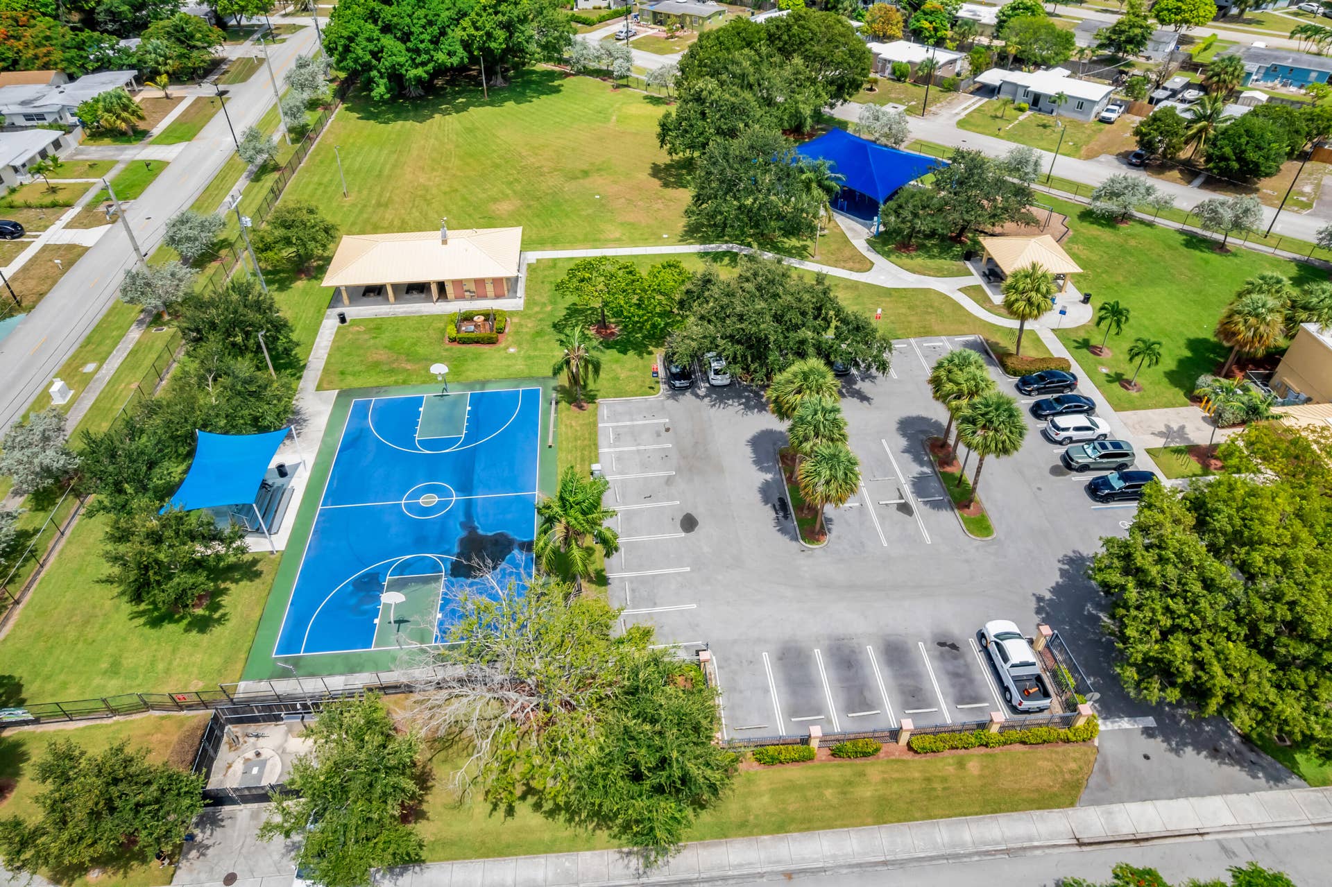 Recreational park grounds featuring a full-size basketball court, covered seating areas, and a paved parking lot with mature palm trees