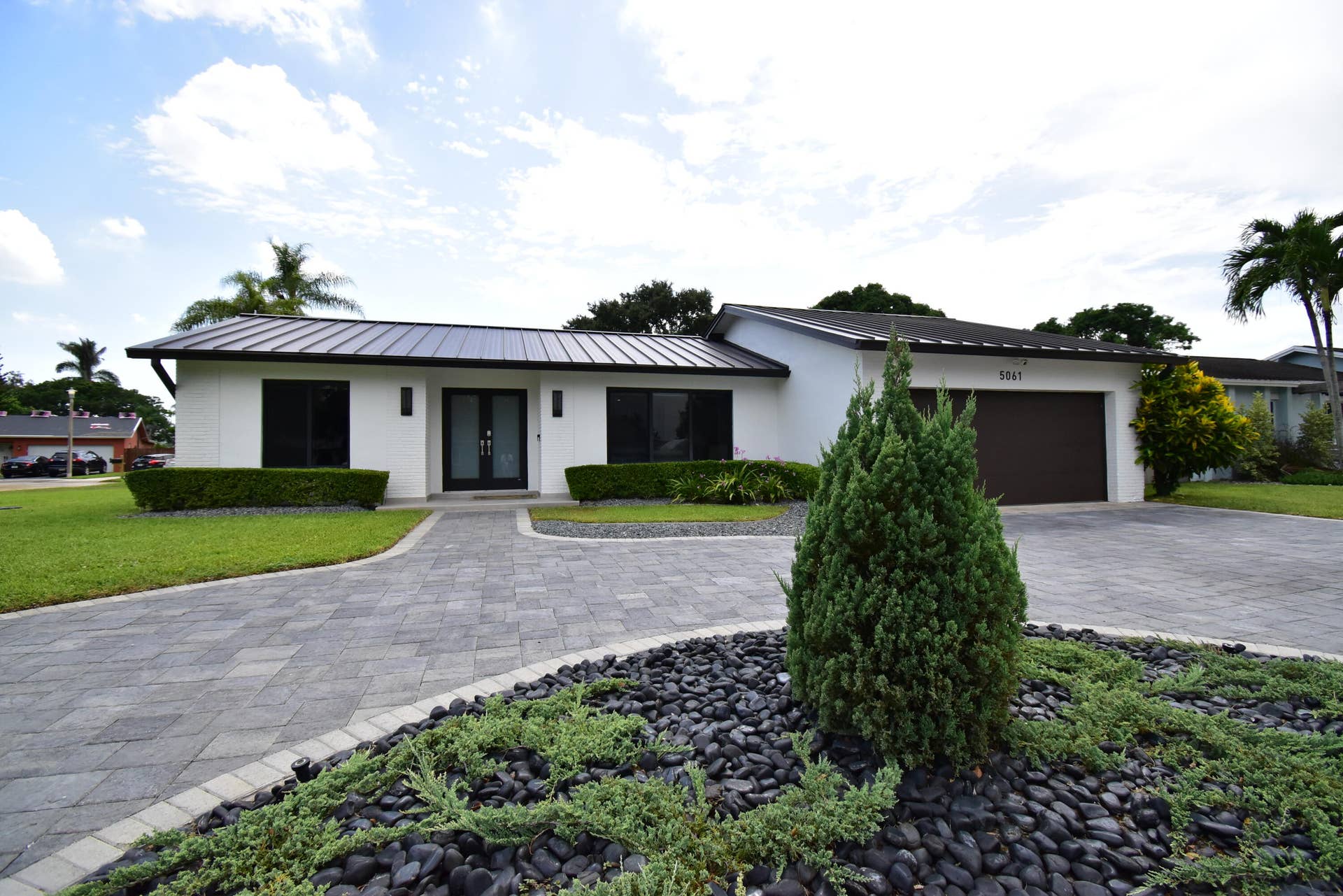 5061 SW 87TH Terrace, Cooper City, FL 33328 Contemporary white exterior featuring a dark metal roof, a paved circular driveway, a dark garage door, and a manicured front lawn with evergreen landscaping