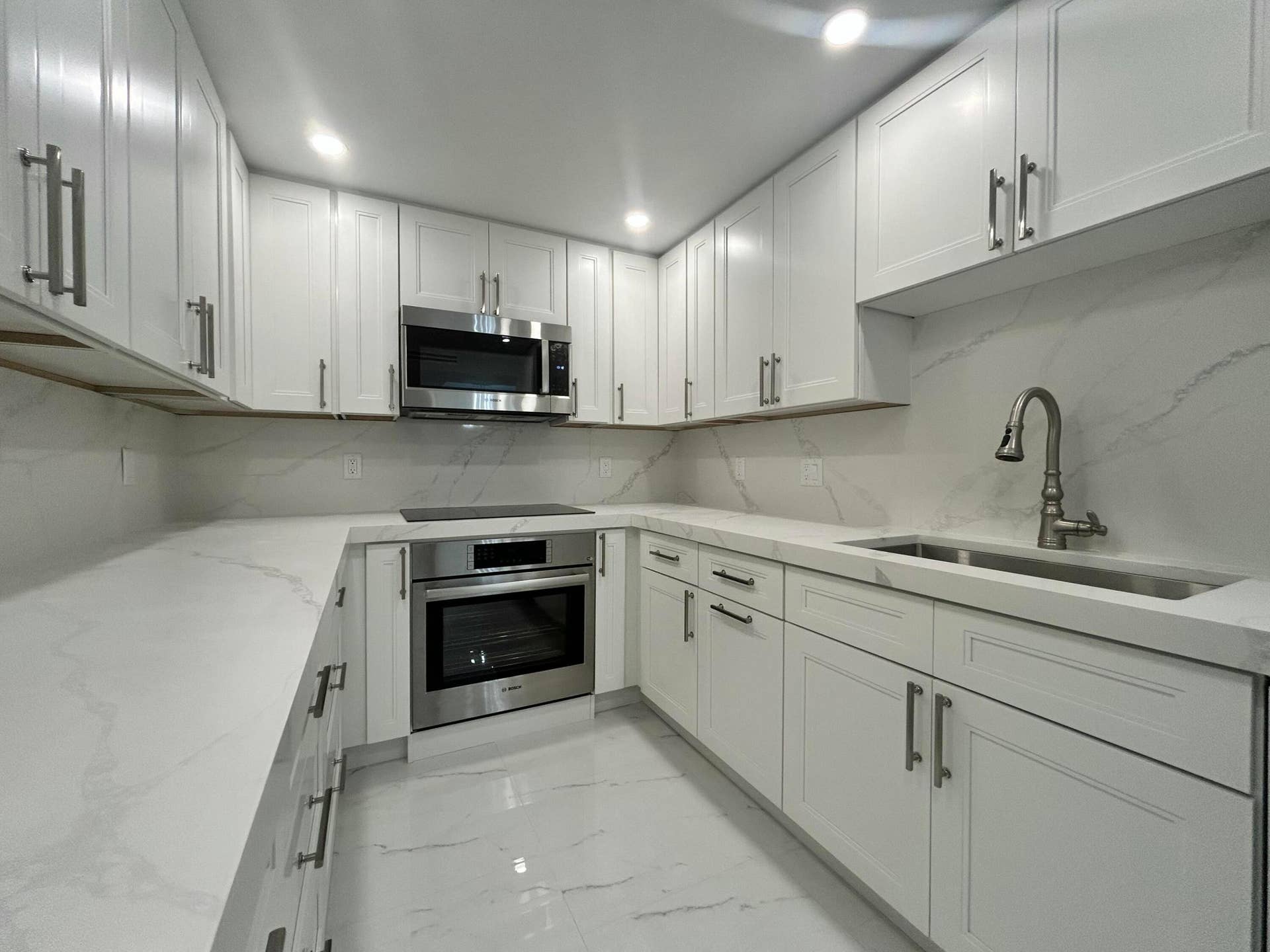Sleek contemporary kitchen featuring white shaker-style cabinetry with brushed metal hardware, white quartz countertops, stainless steel built-in microwave and wall oven, an undermount sink with modern gooseneck faucet, and marble-look tile backsplash