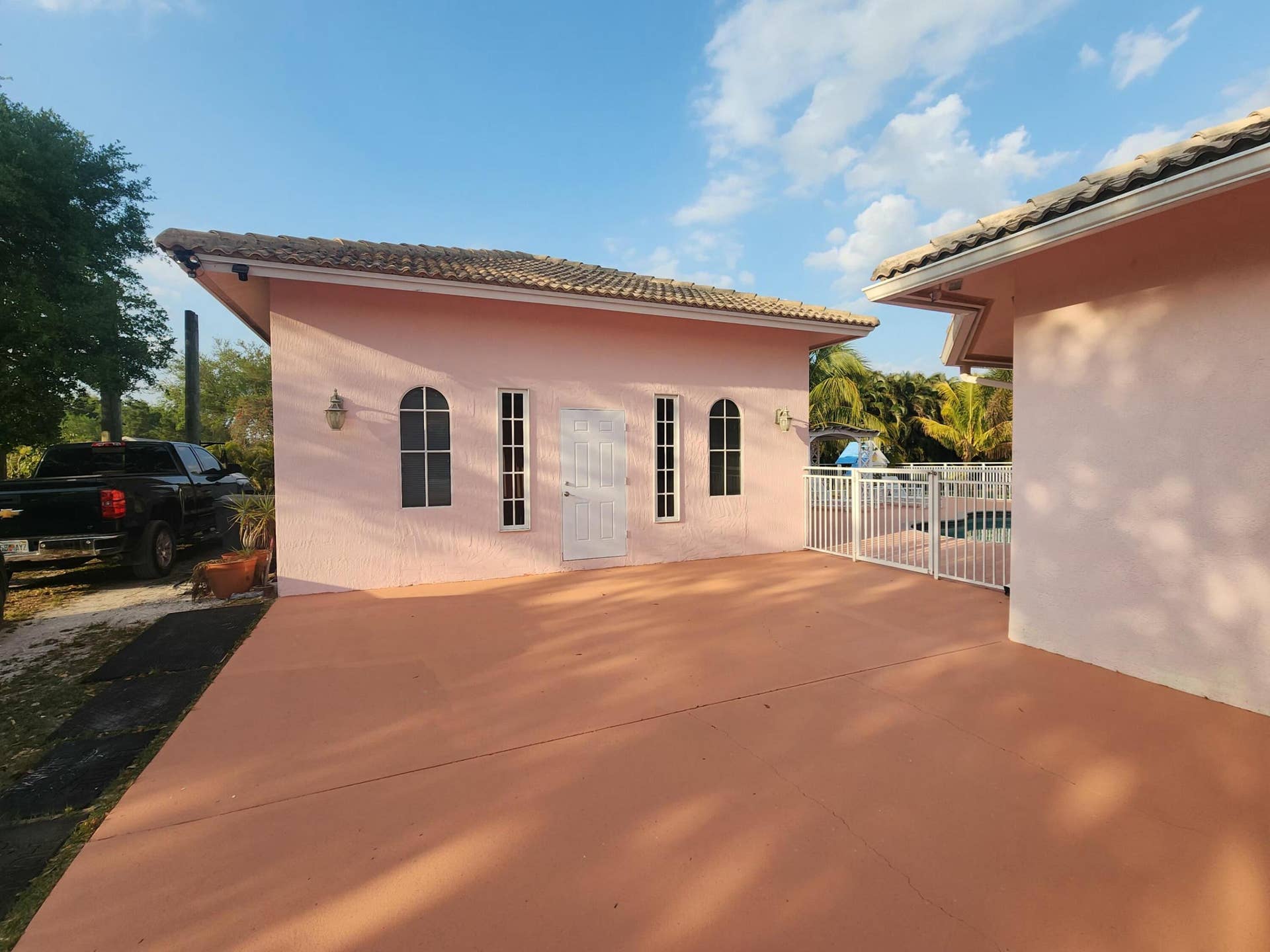 Pink stucco exterior building with a tile roof, arched windows, and a white entrance door, featuring an expansive paved patio area and a white metal fence bordering a pool