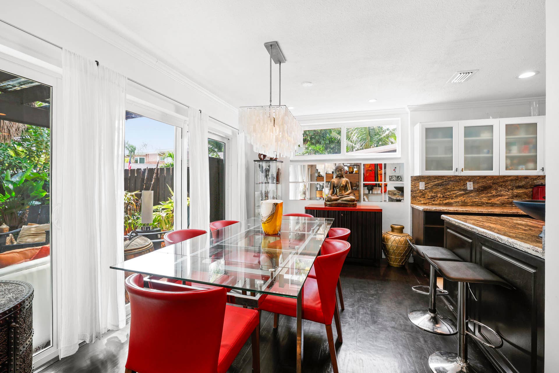 7 Middlesex Drive, Wilton Manors, FL 33305 Contemporary kitchen and dining area showcasing a black island with waterfall countertop edge, stainless steel appliances including a built-in wall oven, white cabinetry with glass-front upper storage, polished concrete flooring, and a glass-top dining table with red upholstered seating