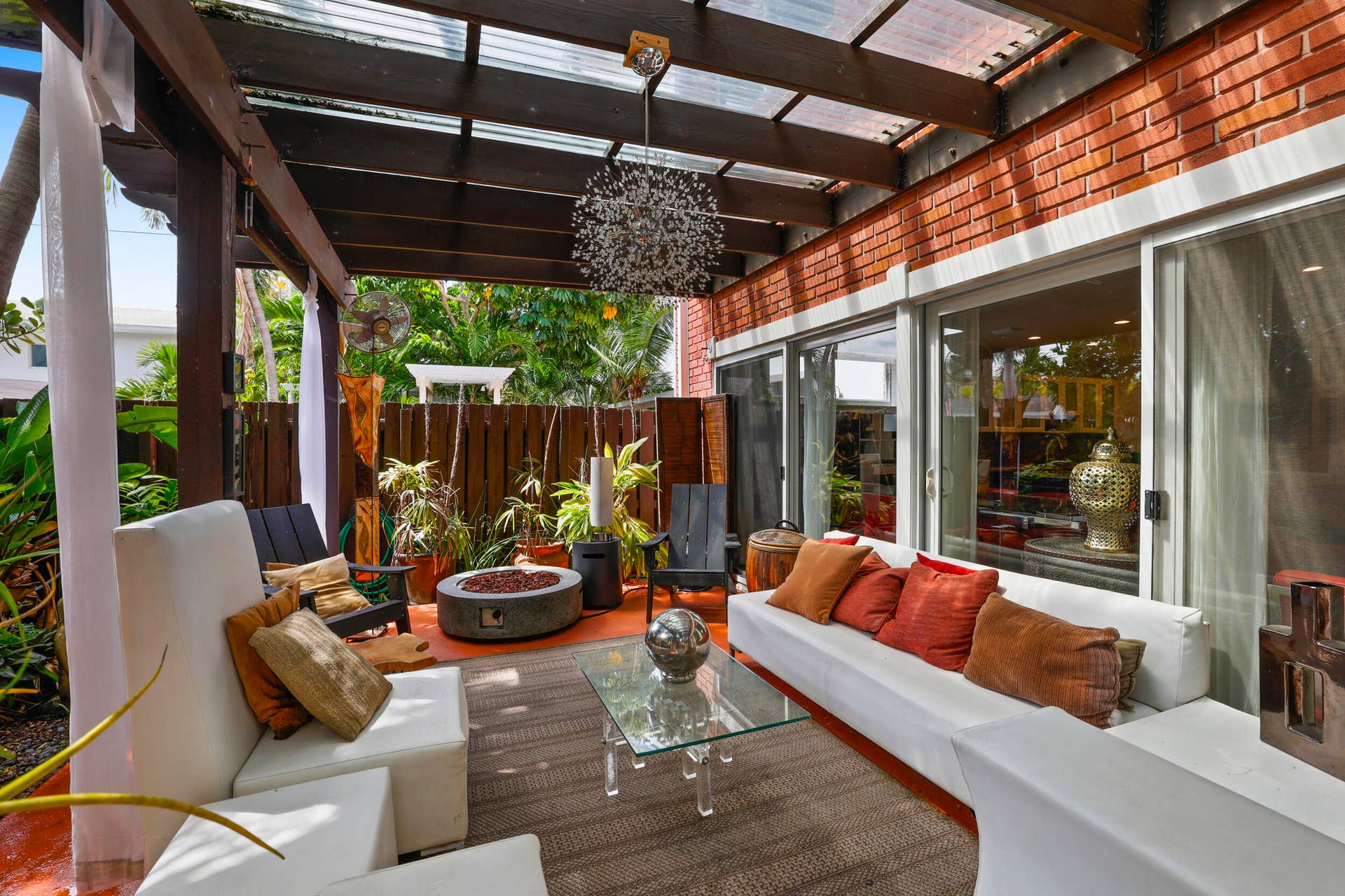 7 Middlesex Drive, Wilton Manors, FL 33305 Covered patio retreat featuring a wood-beam pergola with polycarbonate panels, built-in seating with upholstered cushions and decorative pillows, a fire pit with metal surround, and large multi-panel sliding glass doors opening to the brick exterior