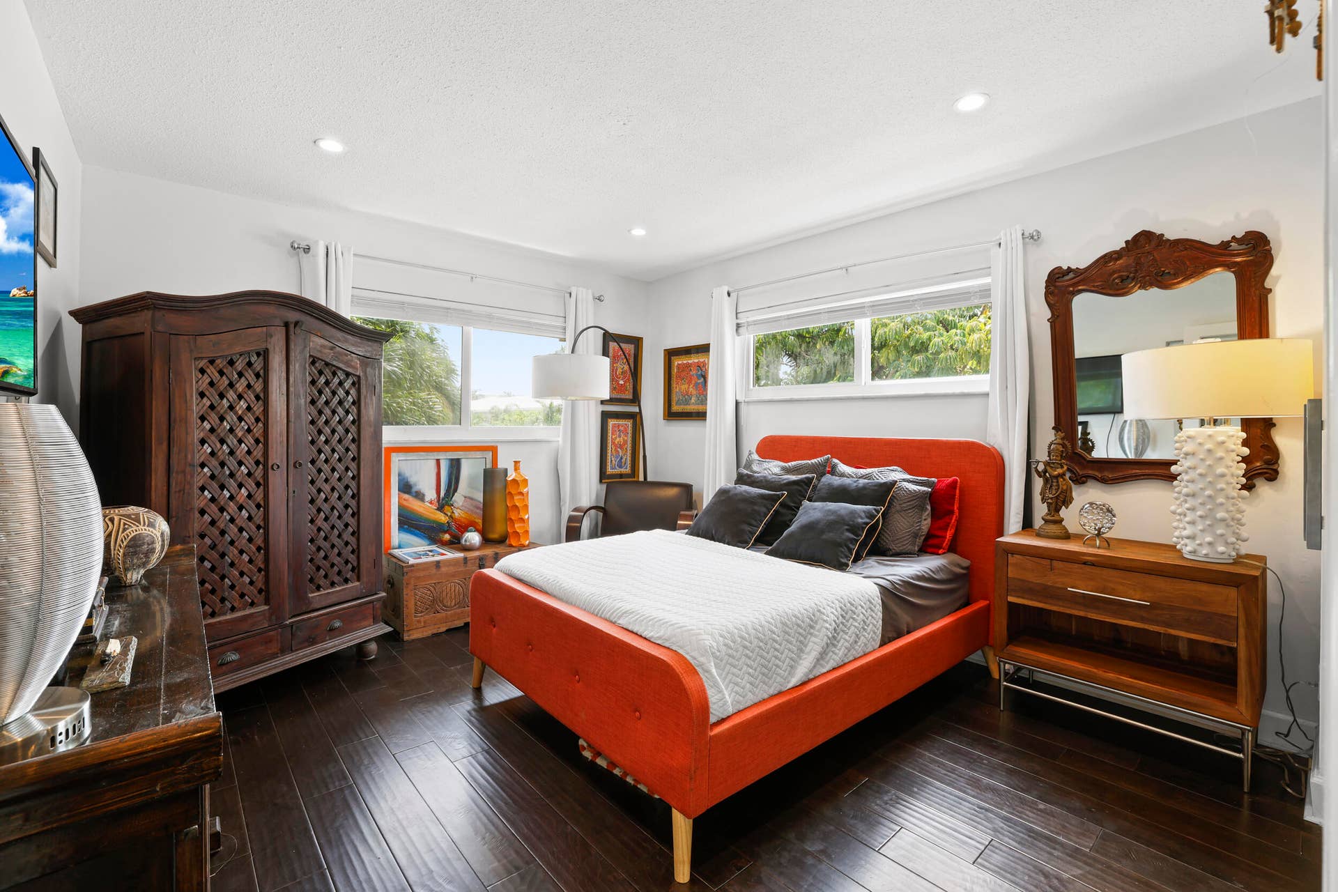 7 Middlesex Drive, Wilton Manors, FL 33305 Spacious primary bedroom featuring a striking upholstered bed frame in burnt orange, dark wood-finish flooring, and multiple large windows flooding the room with natural light
