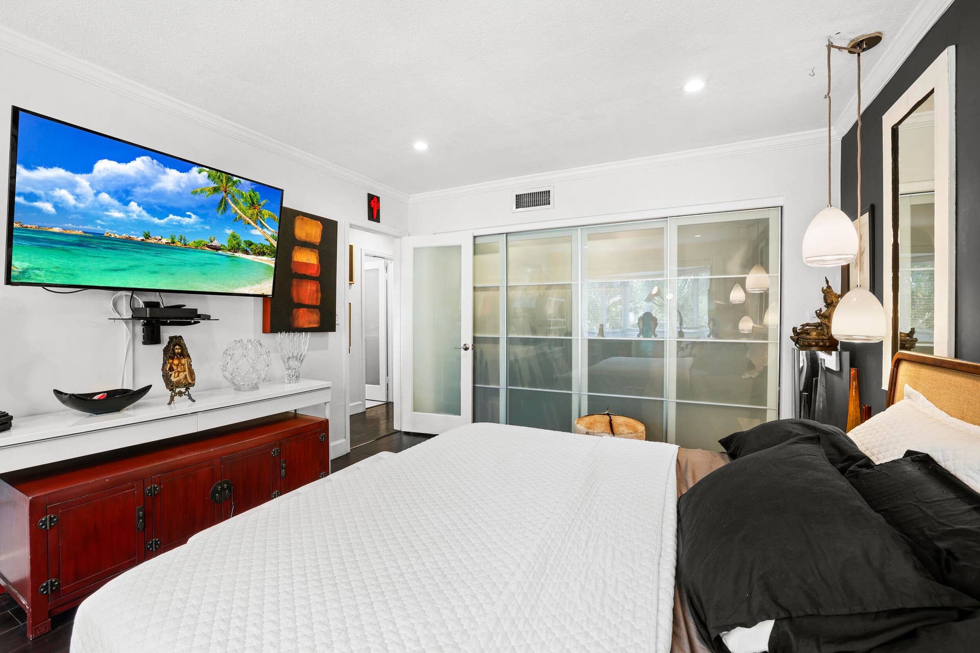 7 Middlesex Drive, Wilton Manors, FL 33305 Spacious primary bedroom featuring a white quilted bed frame, wall-mounted flat-screen television with decorative accent shelving, red lacquered storage chest, and sliding glass doors opening to a private balcony