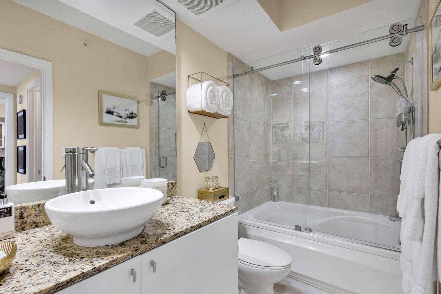 Bathroom featuring a white vessel sink set on a speckled countertop, a mirrored vanity, a commode, and a tub with a sliding glass shower door and tiled surround