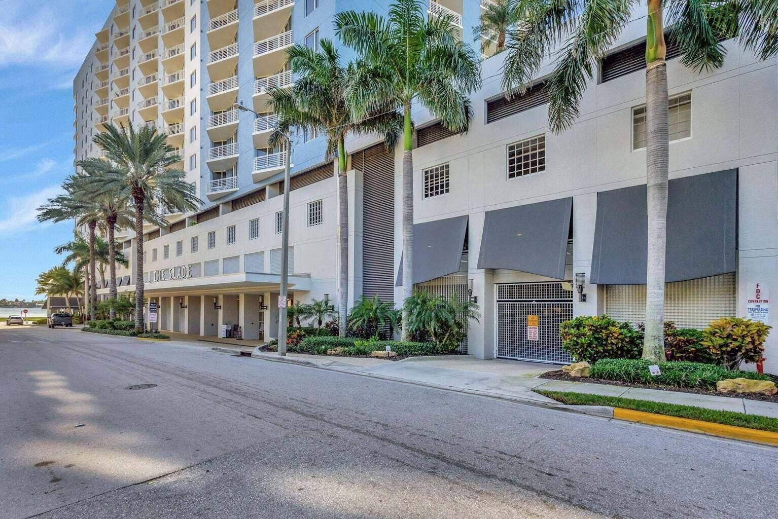 Waterfront high-rise building featuring multiple levels of balconies, ground-floor retail spaces, a gated parking entrance with awning coverage, and tropical landscaping with mature palm trees