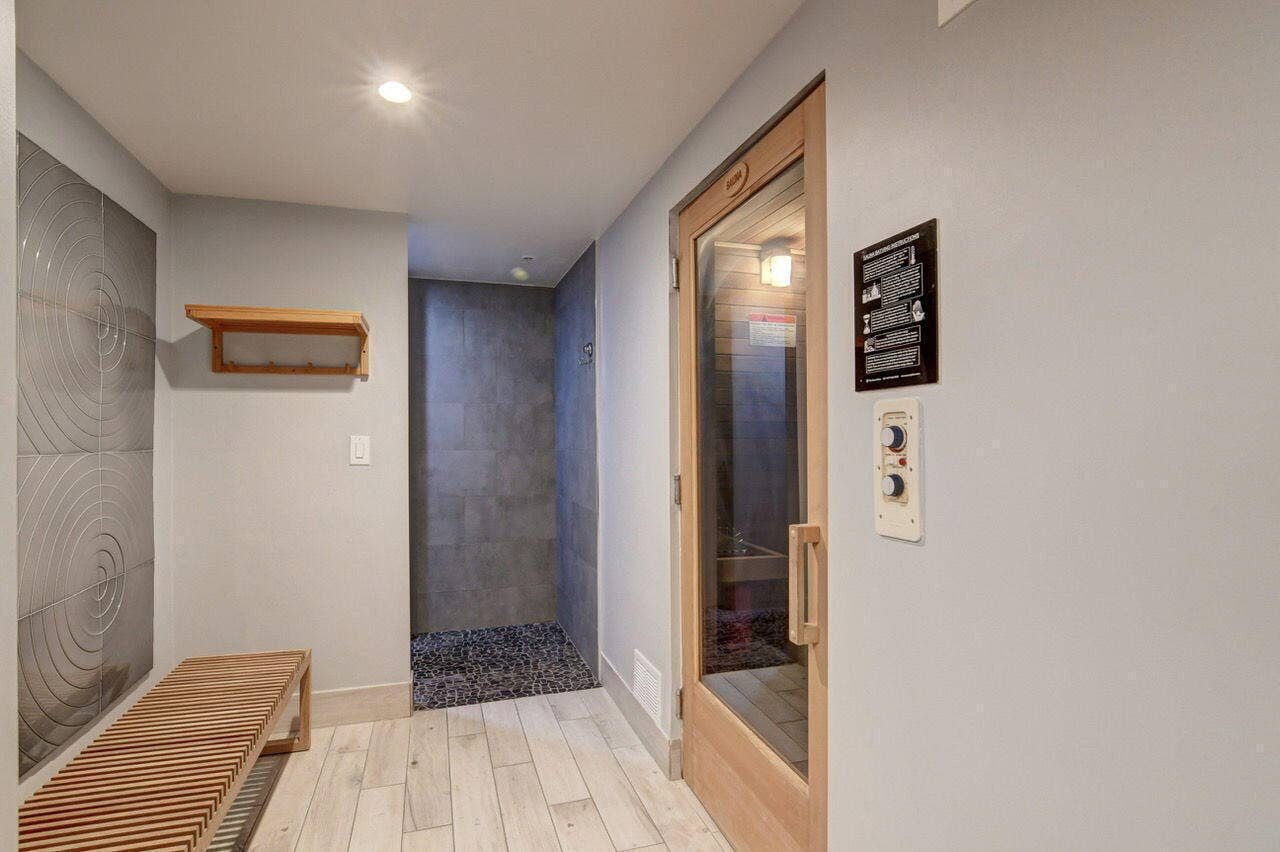 Dedicated sauna room with a clear door and exterior controls, featuring wood-finish flooring, a spa-style shower with dark pebble-stone base and wall tiling, a slatted wood bench, and textured wall panels