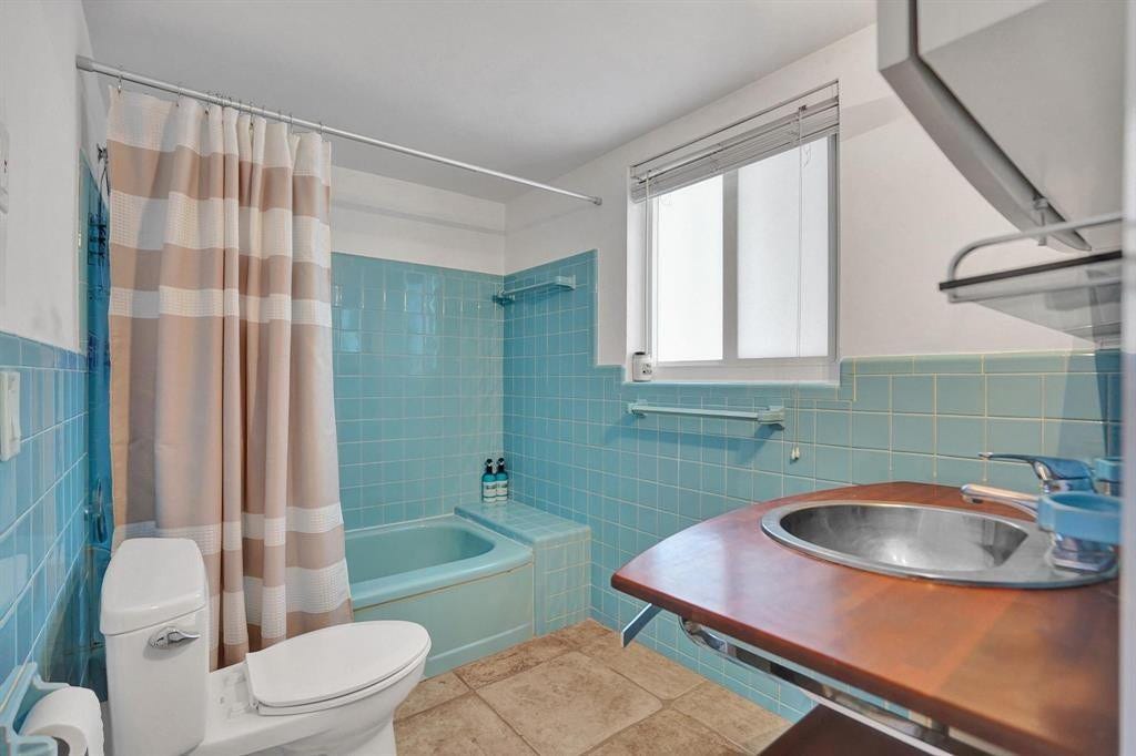2760 YACHT CLUB Boulevard, #10C, Fort Lauderdale, Fort Lauderdale, FL 33304 Bright, updated bathroom featuring turquoise subway tile walls, a soaking tub with shower enclosure and striped curtain, white ceramic toilet, wood-finish vanity with undermount sink, tile flooring, and a window providing natural light