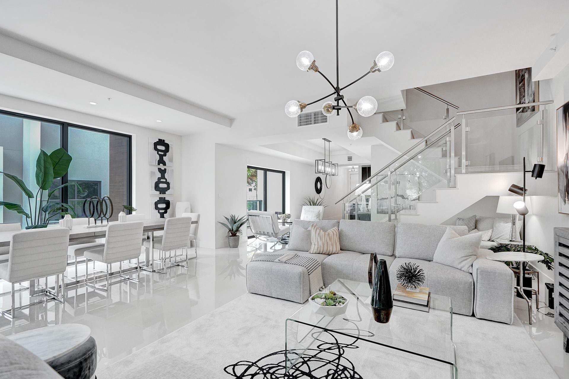 Modern open-concept living space featuring polished light-toned flooring throughout, floor-to-ceiling windows with natural light, a sculptural multi-pendant ceiling fixture, a floating staircase with glass railings, and contemporary furnishings arranged across the expansive great room