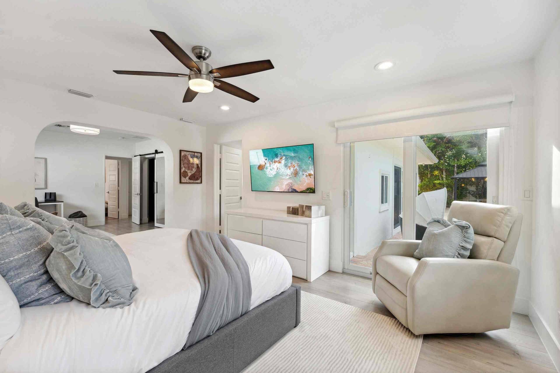 Spacious primary bedroom suite featuring a gray upholstered bed frame, ceiling fan with integrated lighting, ensuite bathroom access, and a comfortable seating area with cream-toned armchair