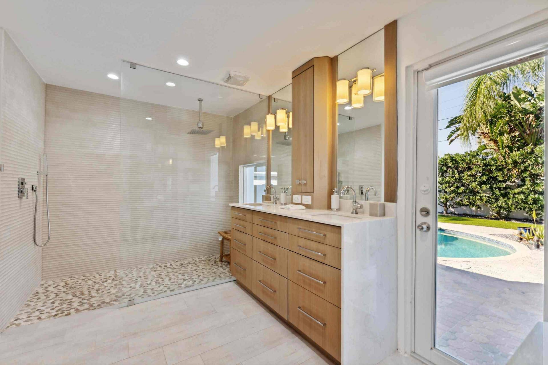 Spa-inspired primary bathroom featuring a spacious walk-in shower with white subway tile surround and pebble stone flooring, custom wood-finish vanity with dual sinks, integrated mirror lighting with warm-toned pendant fixtures, and direct access to the backyard through glass sliding doors