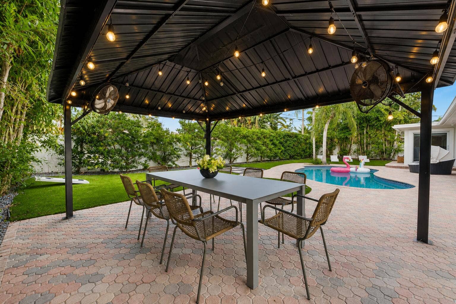 Elegant covered patio featuring a black metal gable roof with integrated pendant lighting and ceiling fans, anchoring a spacious brick-paved entertaining area