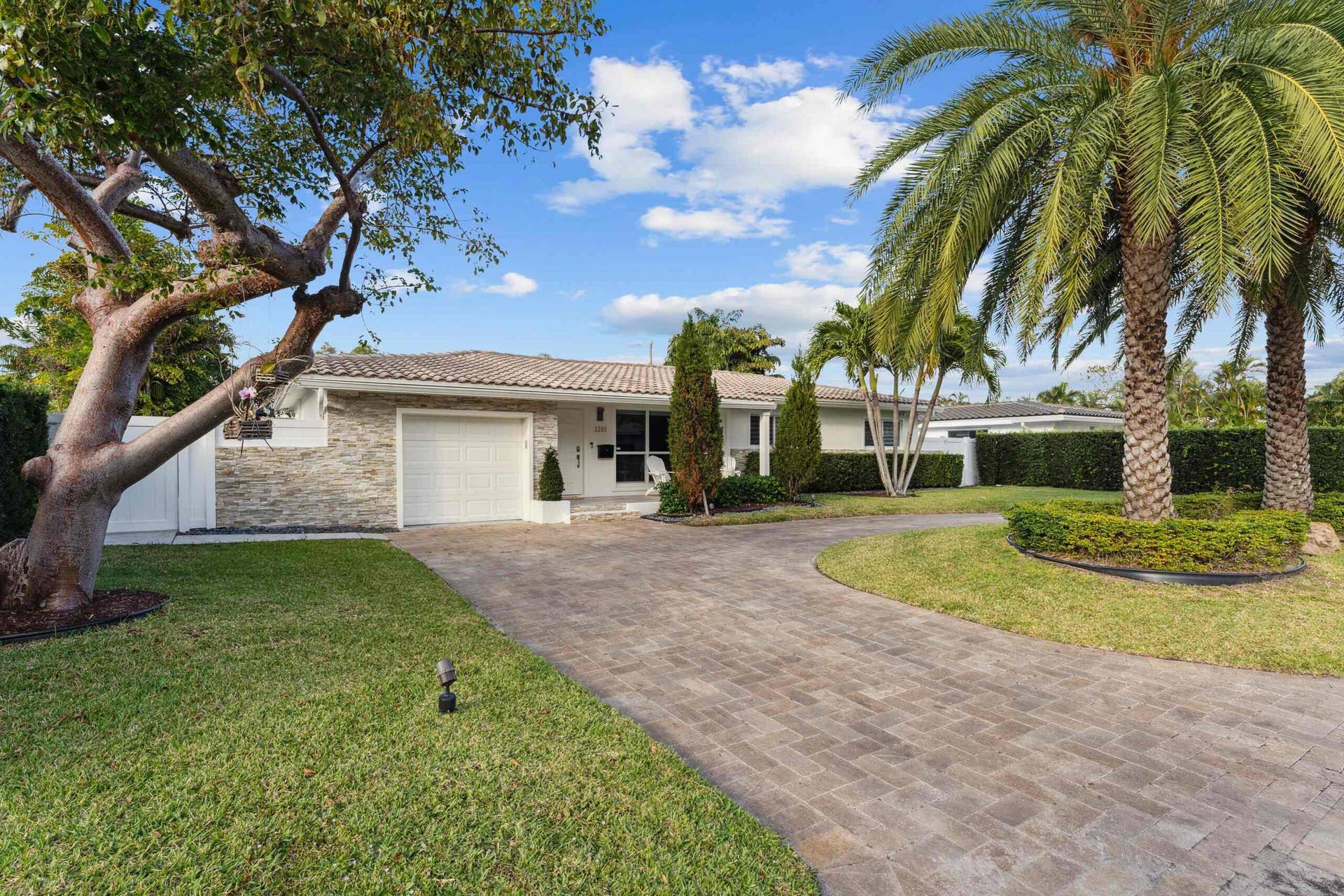 Elegant single-story residence featuring a stone and stucco exterior, wide circular driveway with paver construction, mature landscaping including a striking specimen tree and lush palm plantings, manicured lawn, and professional hedge borders framing the property