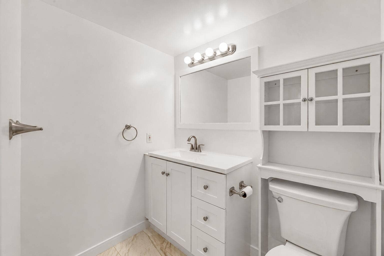 Bright bathroom featuring white cabinetry with multiple drawers and hardware accents, a white vanity with an undermount sink and brushed nickel faucet, framed mirror with integrated lighting, and white glass-front wall cabinet with shelving
