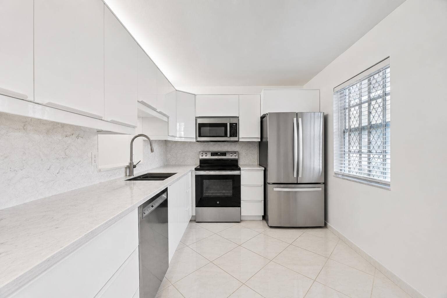 Modern kitchen featuring stainless steel appliances, white cabinetry with sleek hardware, marble-finish backsplash, tile flooring, and abundant natural light from a large window