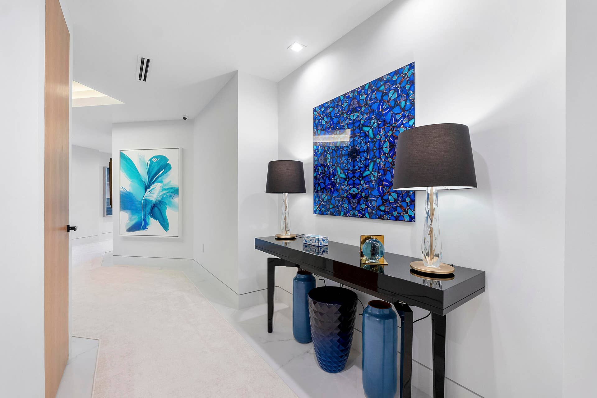 1700 S OCEAN Boulevard, #PH B-D, Lauderdale By The Sea, FL 33062 A Private Passage Through Art & Design The softly carpeted En-Suite Gallery creates an elegant transition into the penthouses private bedroom wing. Beginning beside the vibrant Mandarin Room, the gallery gently winds toward the Primary and Rosewood Suites, unfolding through a curated collection of striking sapphire-blue artwork and Asian-inspired botanical accents. Along the way, lush green imagery and sculptural details create a beautiful rhythm of color and discovery, while a discreetly concealed butlers pantry, second laundry and refreshment bar virtually disappear within the passage. In this photograph, the dramatic sapphire artwork immediately draws the eye down the gallery, while the glimpse of vivid botanical art beyond hints at the rooms still to be discovered.