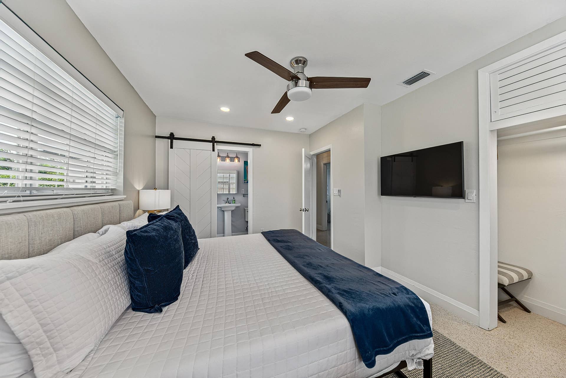 Spacious primary bedroom featuring a wood-finish ceiling fan, recessed lighting, dual windows with plantation shutters, wall-mounted flat-screen television, and direct access to an ensuite bathroom
