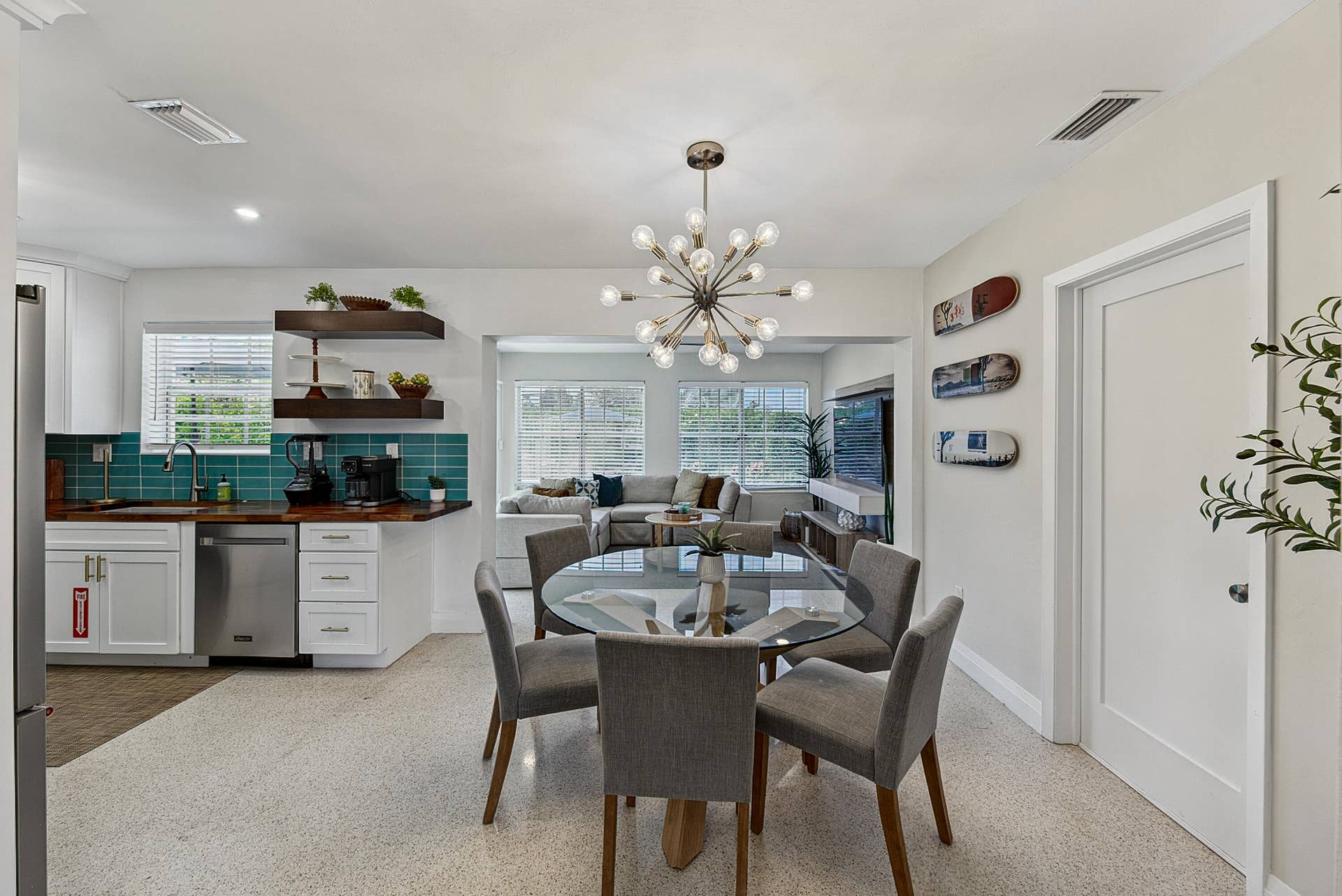 Open-concept dining area with a contemporary chandelier, durable terrazzo flooring, and a kitchen featuring white cabinetry, a teal tile backsplash, and wood-finish countertops