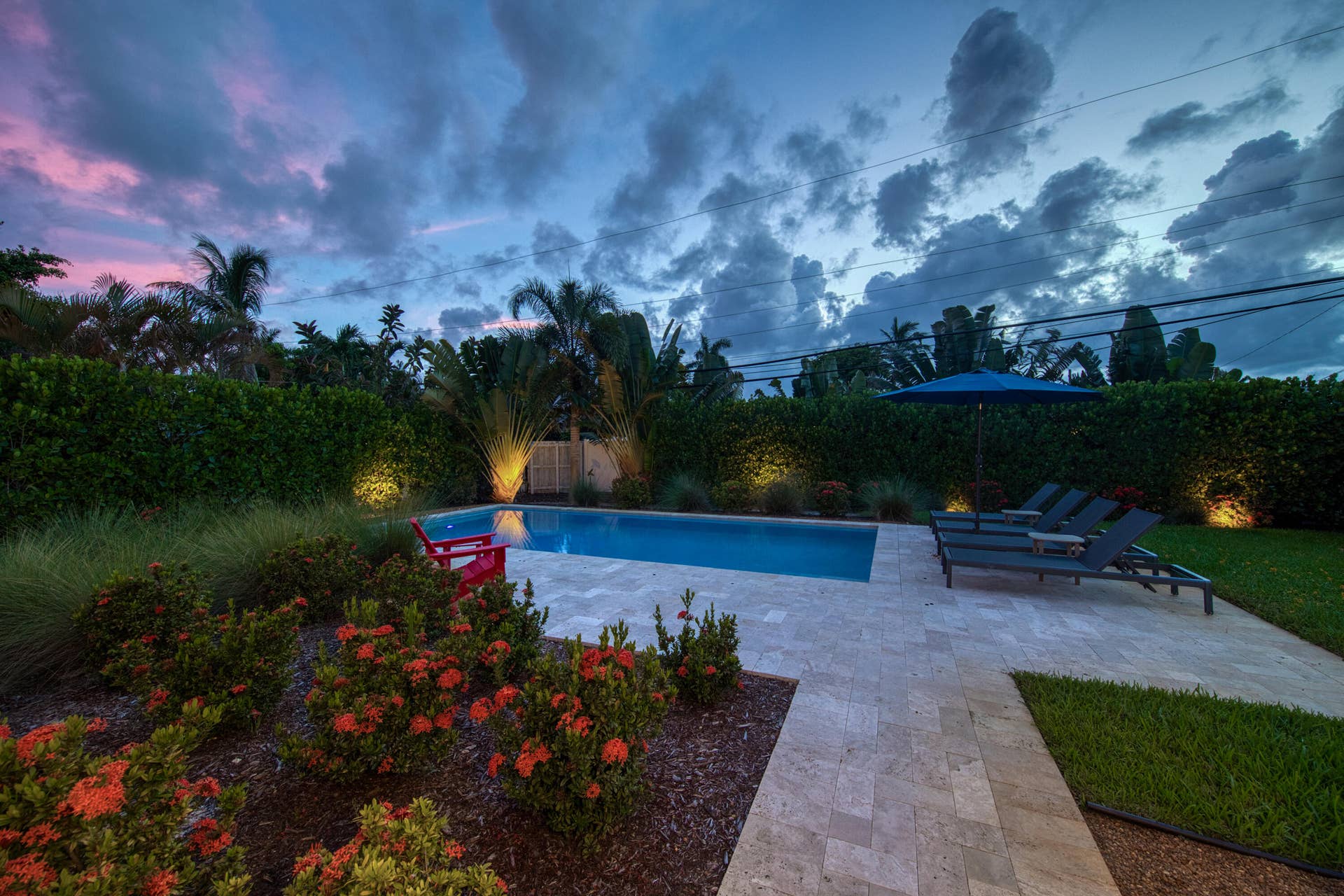 Expansive backyard featuring a rectangular swimming pool, light-toned paved patio, manicured lawn, and lush landscaping with accent lighting