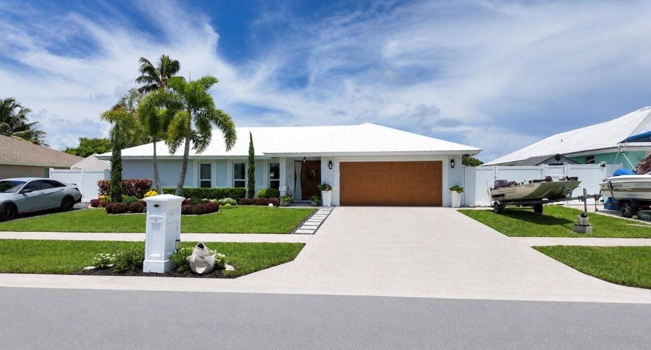 1201 SW 25TH Avenue, Boynton Beach, FL 33426 Lush green lawn with tropical landscaping, a white metal roof, and a wood-finish garage door