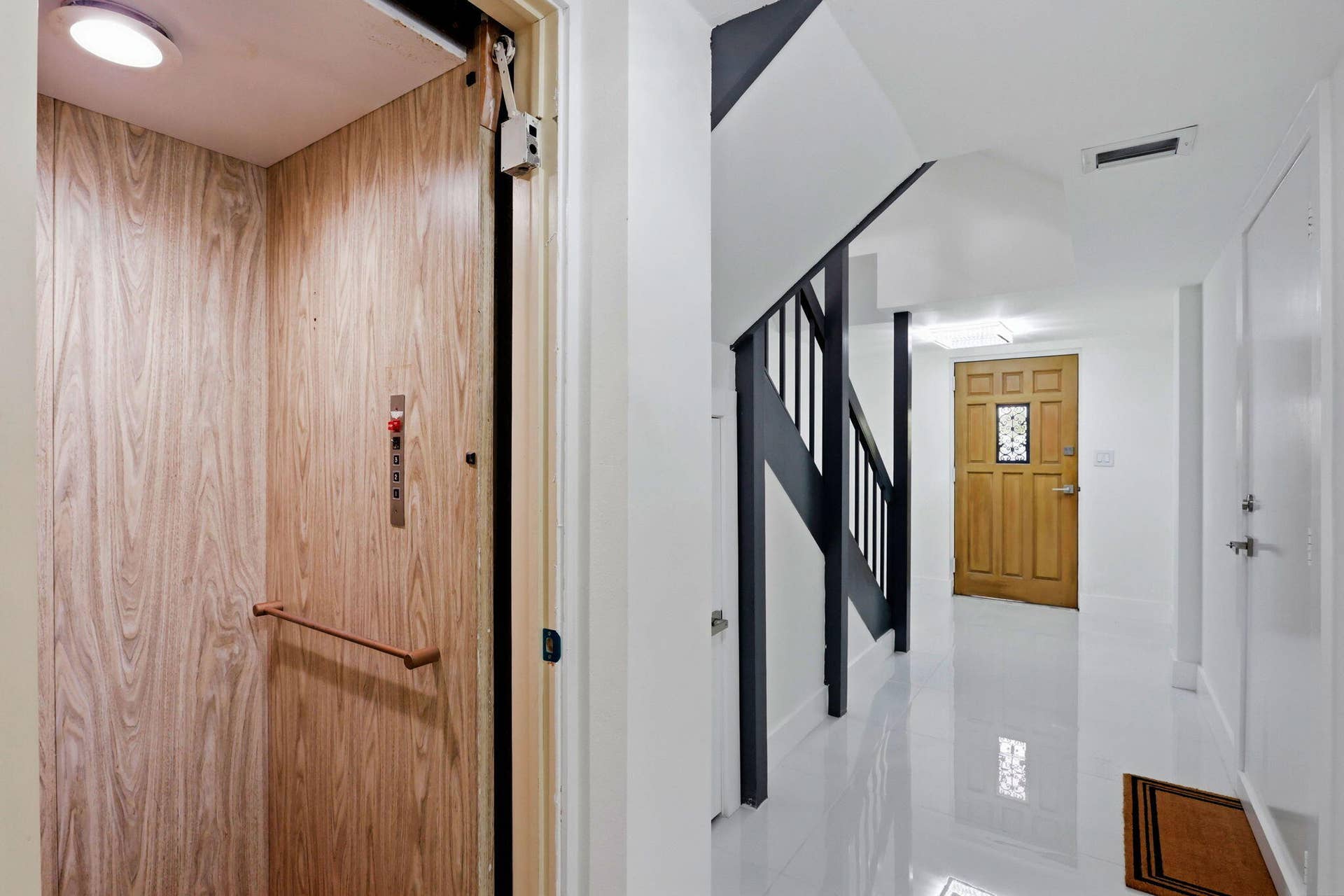 1222 Hillsboro Mile, #15, Hillsboro Beach, FL 33062 Wood-finish interior elevator with control panel and handrail, alongside a hallway featuring high-gloss flooring, a dark-stained staircase, and a paneled wooden door with a decorative glass insert