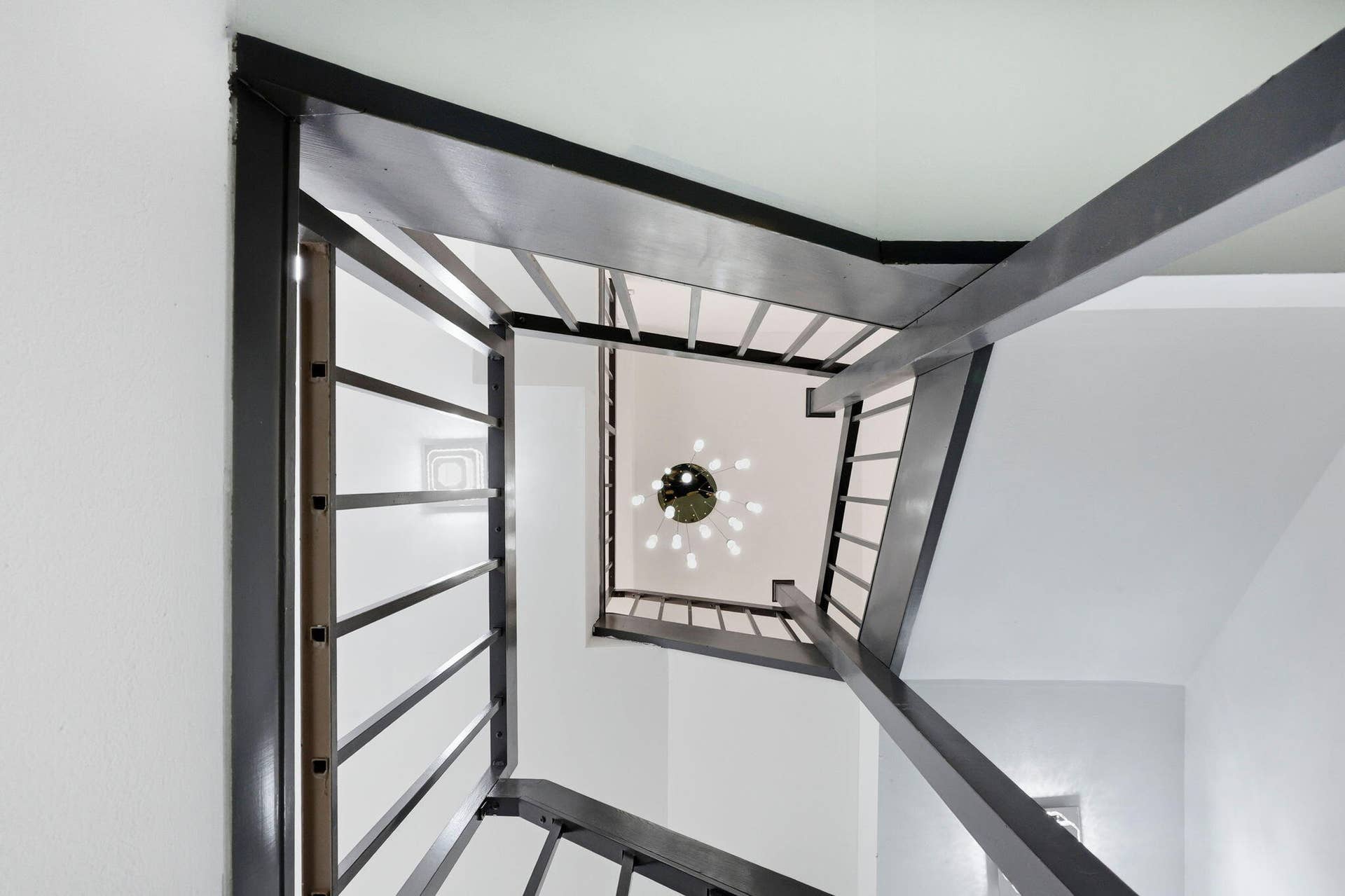 Architectural staircase with metal railings, white walls, and a multi-bulb ceiling fixture