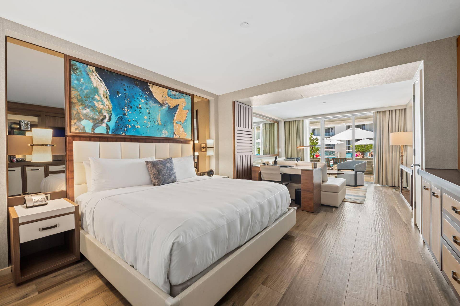 Spacious bedroom suite featuring a king bed with upholstered headboard and integrated bedside lighting, wood-finish flooring throughout, and floor-to-ceiling windows opening to a private sitting area