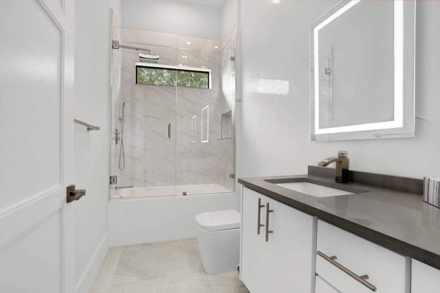 2517 Oceanview Avenue E, Delray Beach, Delray Beach, FL 33444 Bathroom featuring a glass-enclosed shower-bathtub combination, floating vanity with an integrated sink, rectangular illuminated mirror, and a horizontal window
