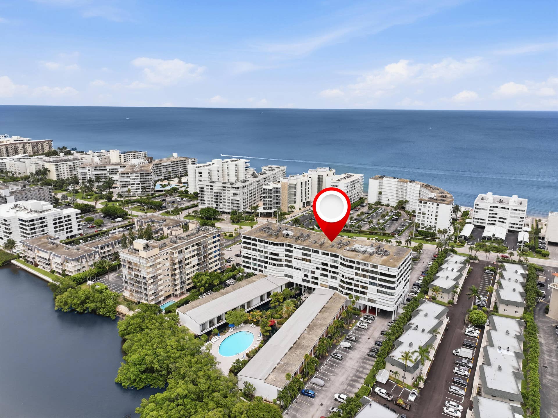 3589 S Ocean Boulevard, #808, South Palm Beach, FL 33480 Ocean to Intracoastal multi-story building featuring a white exterior with horizontal architectural elements, surrounded by extensive parking areas, mature landscaping, and a nearby water body