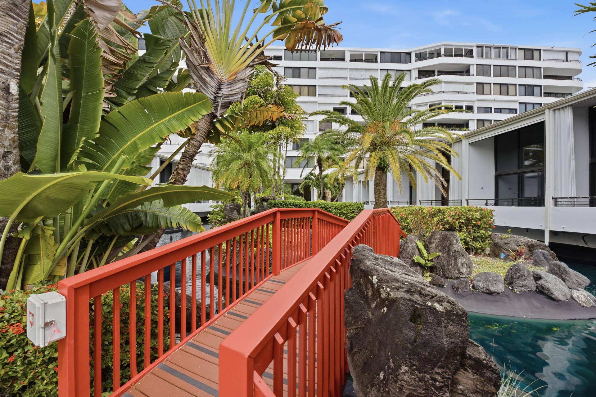 3589 S Ocean Boulevard, #808, South Palm Beach, FL 33480 Decorative red pedestrian bridge over a water feature, surrounded by tropical landscaping, with a multi-story building in the background