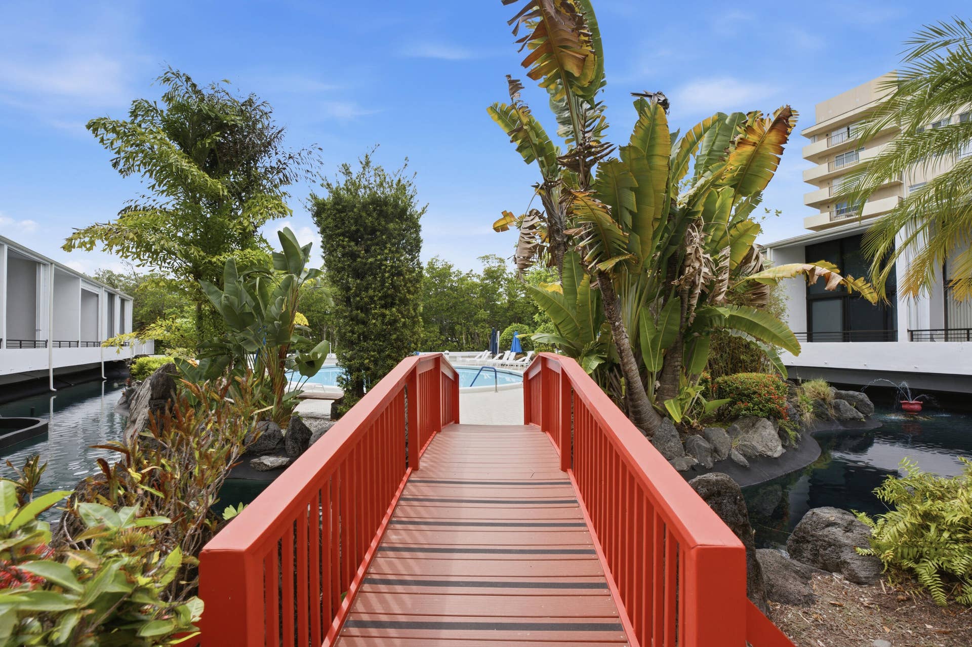 3589 S Ocean Boulevard, #808, South Palm Beach, FL 33480 Red wooden pedestrian bridge over a dark water feature, surrounded by dense tropical foliage and rock landscaping