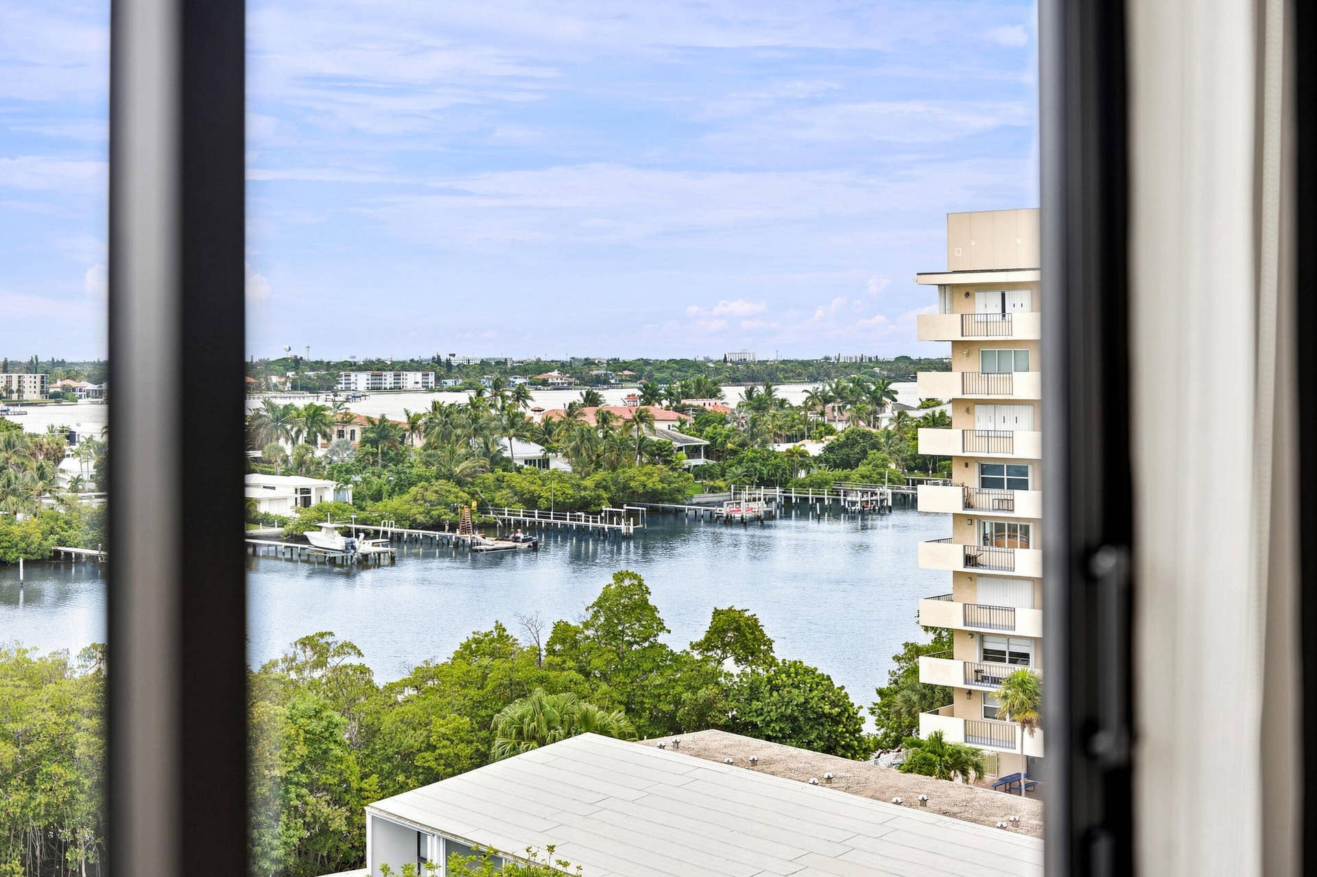 3589 S Ocean Boulevard, #808, South Palm Beach, FL 33480 Expansive water vista framed by an architectural window, featuring a waterway, distant residential properties, lush green landscaping, and a multi-story building with multiple balconies